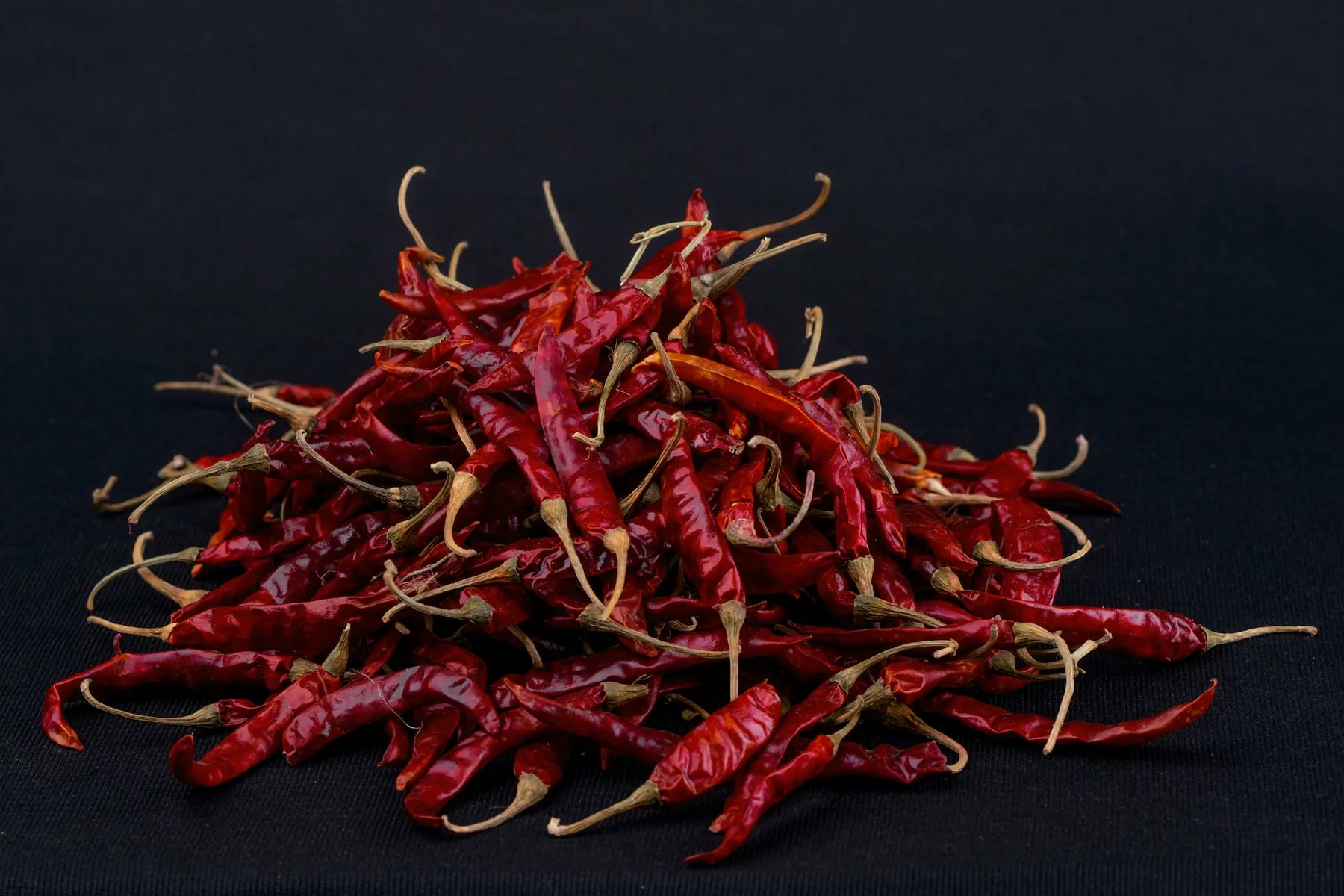 A pile of dried red chillies with stems on a dark surface